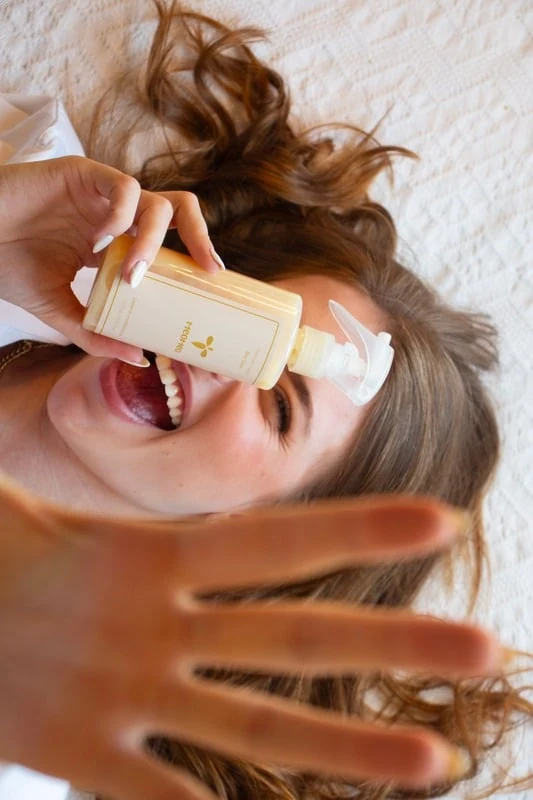 Sara deitada em uma manta clara, sorrindo e segurando um produto de cabelo ReoNeo