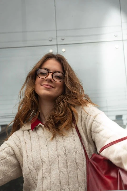 Sara usando óculos de grau, tricô claro e bolsa vermelha, em um retrato na rua