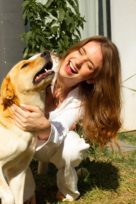 Sara sorrindo ao lado de um cachorro, em um jardim ensolarado